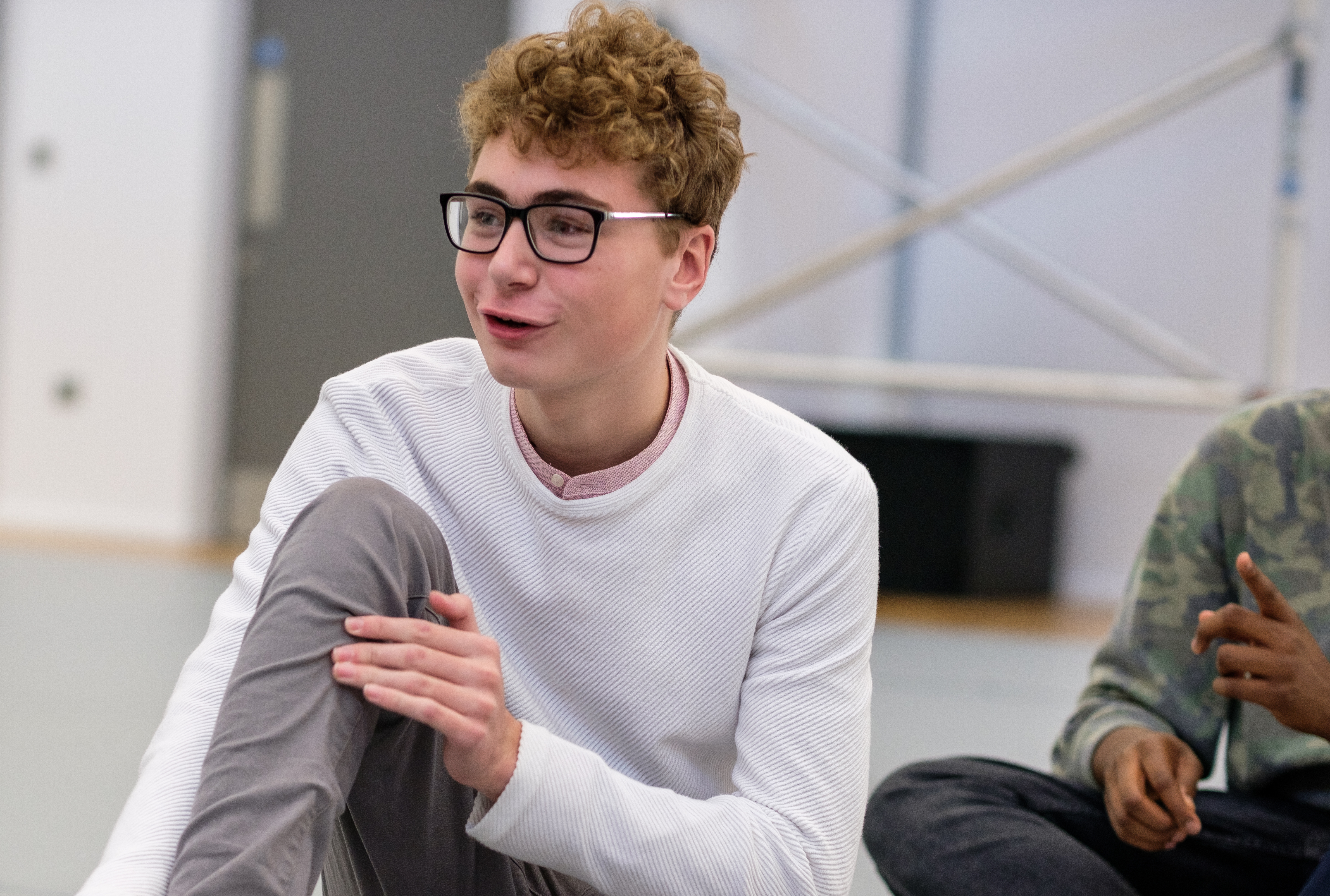 A young man in white jumper and glasses sits on the floor